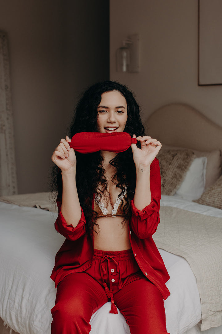 Model sitting on bed in red pyjamas holding a red bamboo sleep mask in front of her face.