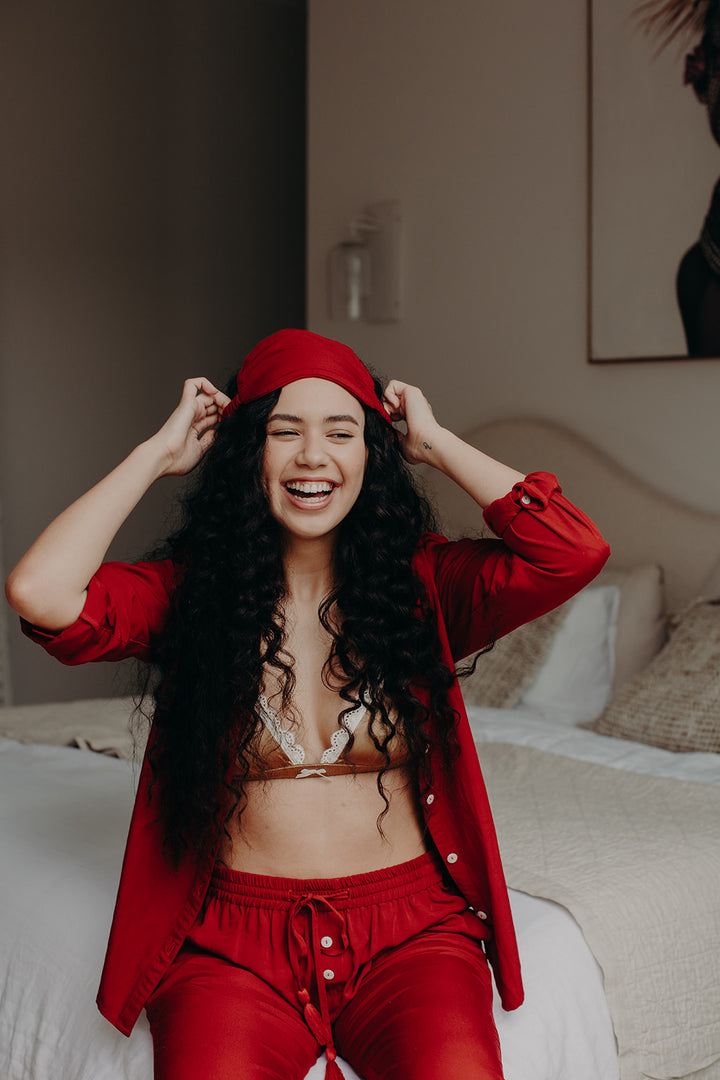 Model on bed wearing red pyjamas, smiling while holding a black bamboo sleep mask in front of her face