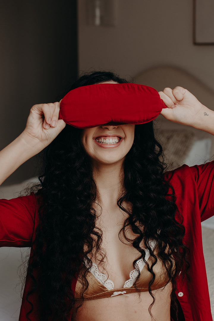 Model in red pyjamas lifting a red bamboo sleep mask above her head with a playful expression.