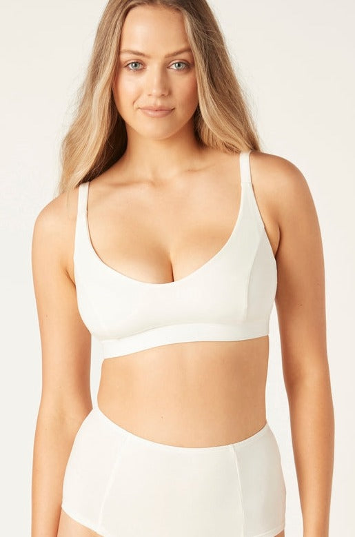 A model wearing an off-white, full cup crop-style bra with adjustable shoulder straps and a racerback option, made from superior organic cotton stretch fabric.