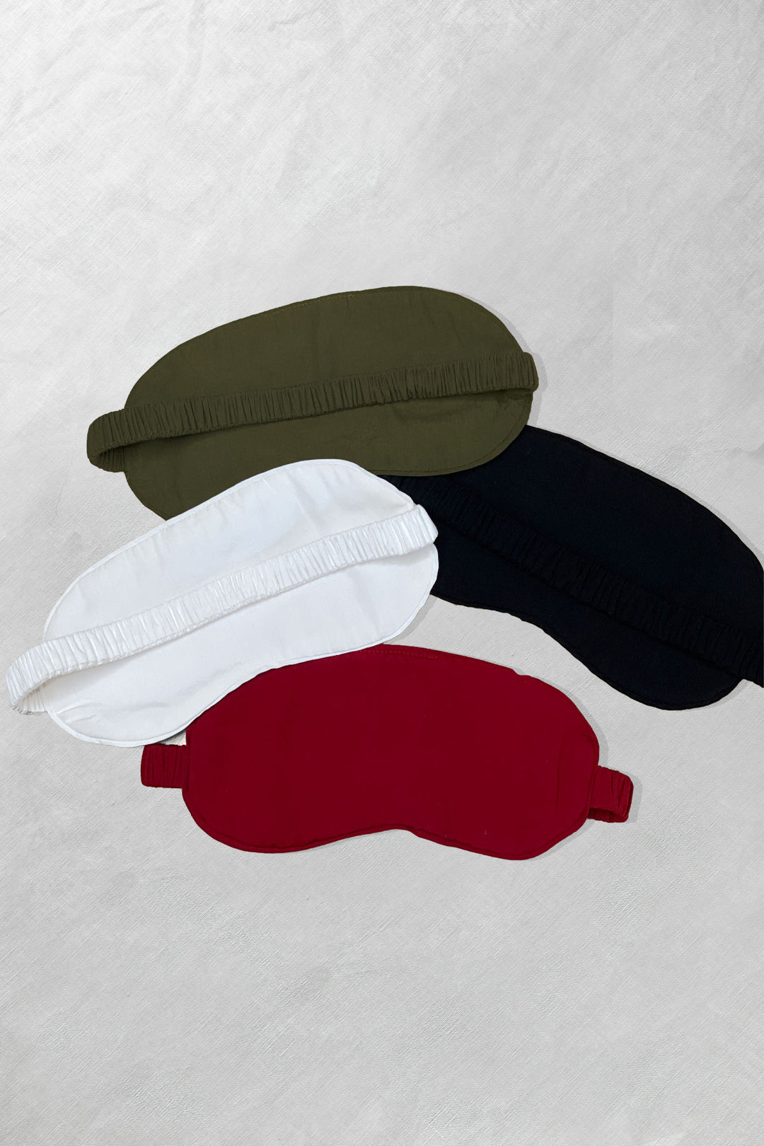 Flat lay of four bamboo sleep masks in olive, white, red, and black, arranged on a light surface.