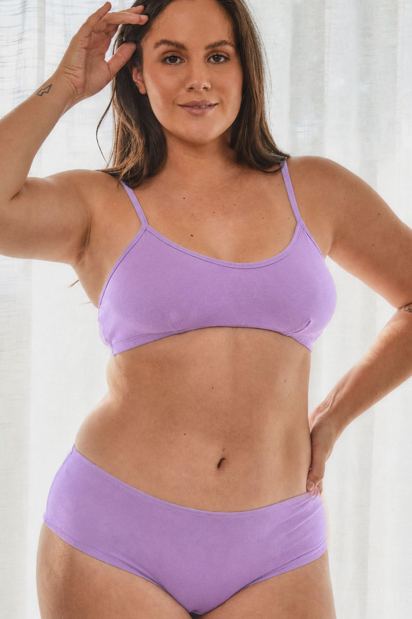 A woman modeling a light purple/lavender two-piece bikini set against a soft white curtain background.