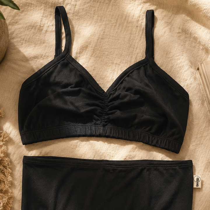 A black two-piece lingerie set laid flat on a beige textured fabric background, featuring a ruched underwire bra top and matching high-waisted briefs.