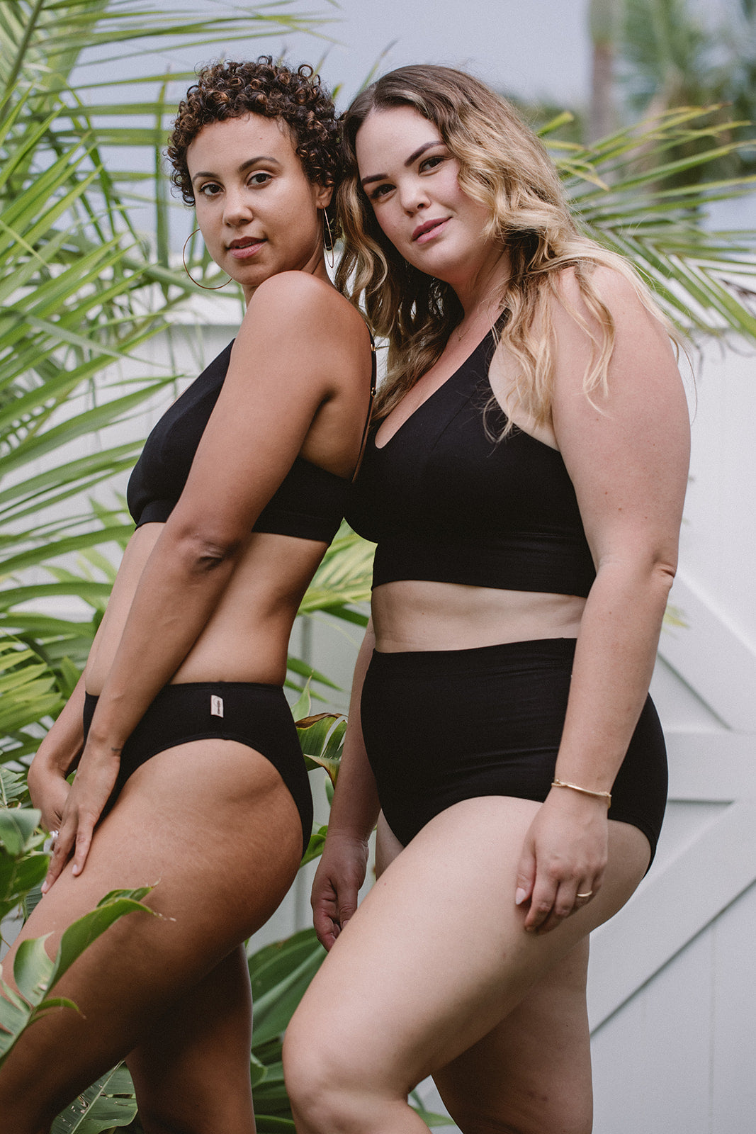 Two women wearing black underwear standing outdoors with greenery in the background