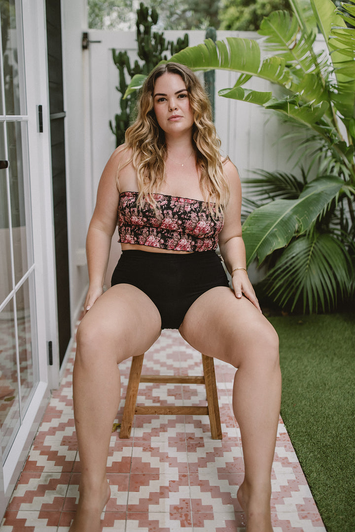 Woman wearing strapless bra sitting on a stool outdoors with plants in the background