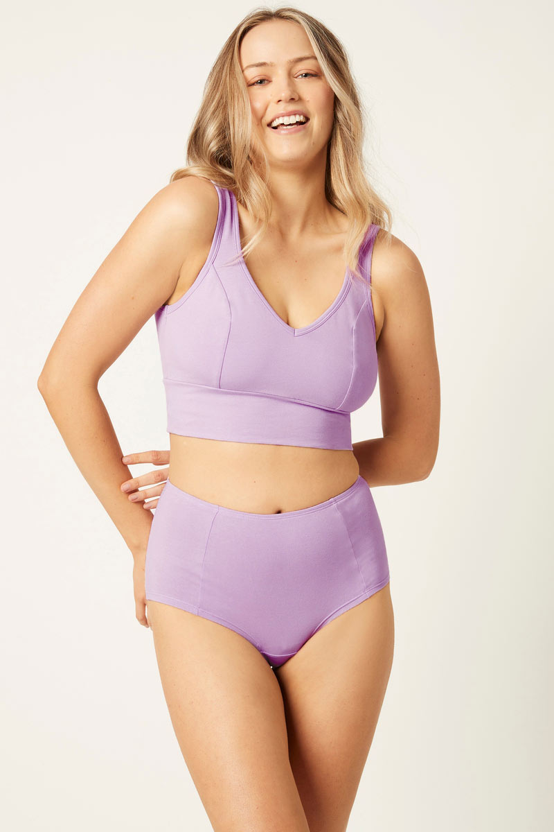 A woman modeling a matching lavender two-piece underwear or lounge set with a scoop-neck crop top and high-waisted briefs, photographed against a plain light background.