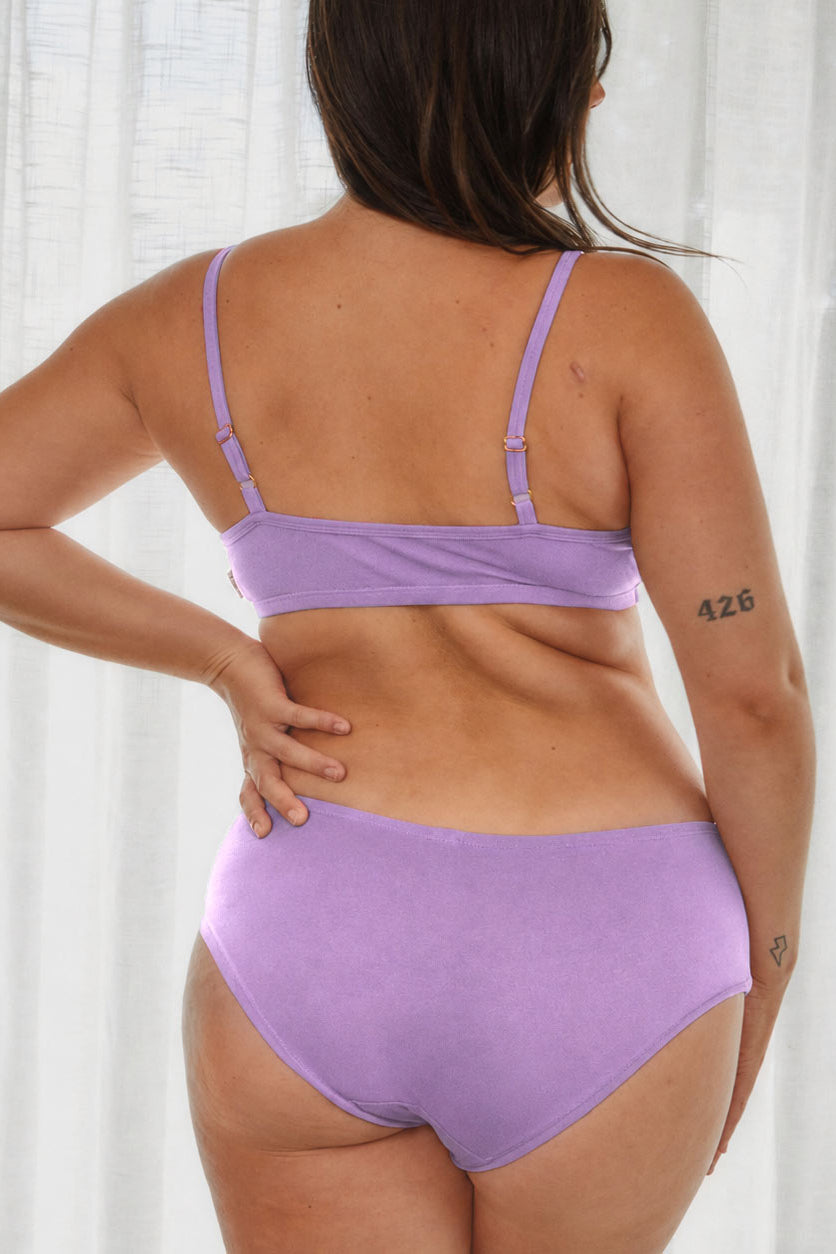 A woman shown from the back wearing a light purple/lavender two-piece bikini set against a soft white curtain background.