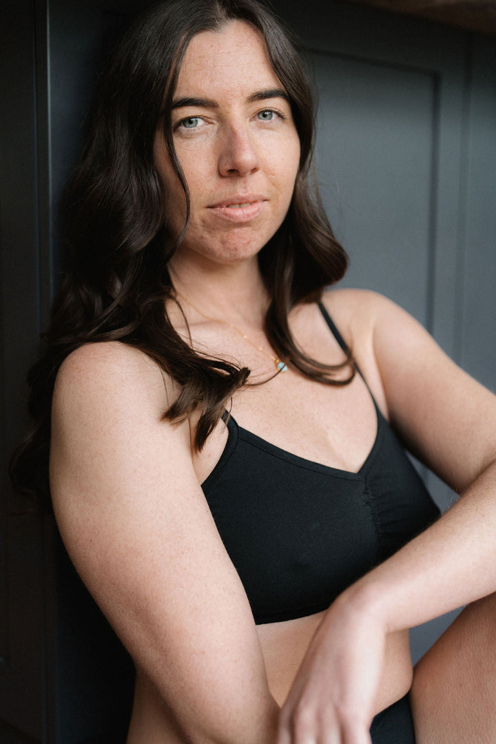 A woman with long dark brown hair wearing a black camisole top, posing against a dark gray paneled background.