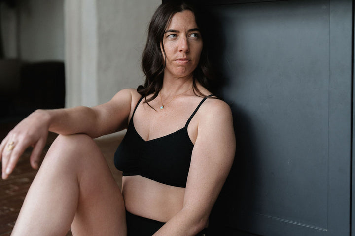 A woman with dark hair sits against a dark gray wall wearing a black bralette and matching bottoms, looking off-camera with a neutral expression.