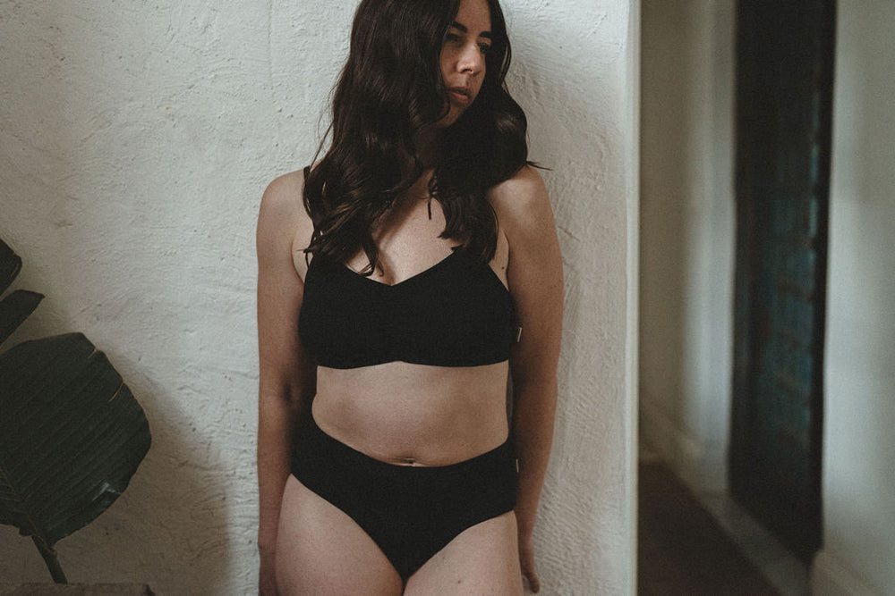 A woman with long dark hair wearing a black organic cotton two-piece underwear set, standing against a textured off-white wall in an indoor setting.