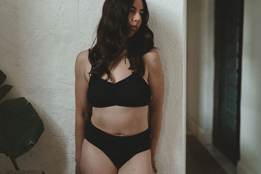 A woman with long dark hair wearing a black organic cotton two-piece underwear set, standing against a textured off-white wall in an indoor setting.
