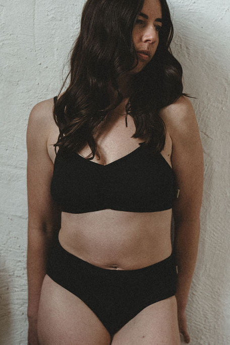 A woman wearing a black two-piece lingerie set, consisting of a organic cotton bralette and high-waisted bottoms, standing against a textured off-white wall.