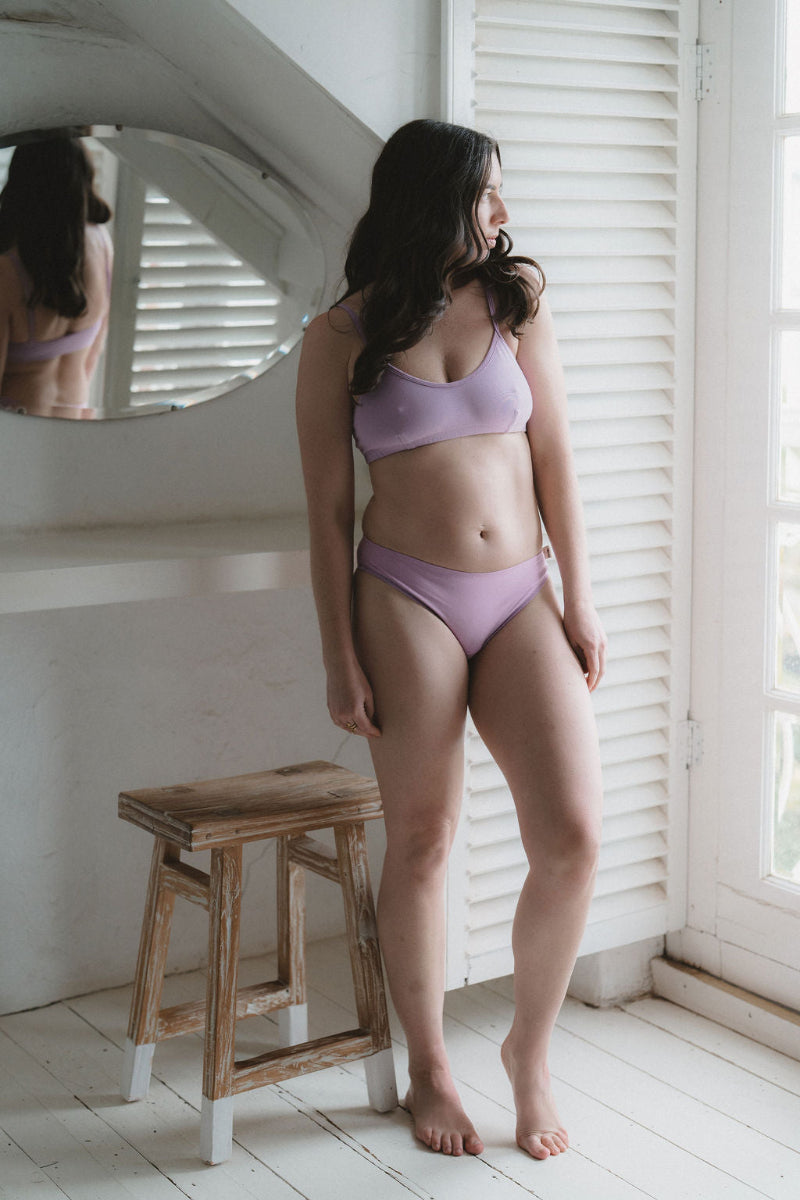 A woman stands barefoot in a bright room wearing a light purple/lavender two-piece lingerie set, with a wooden stool and window shutters in the background.